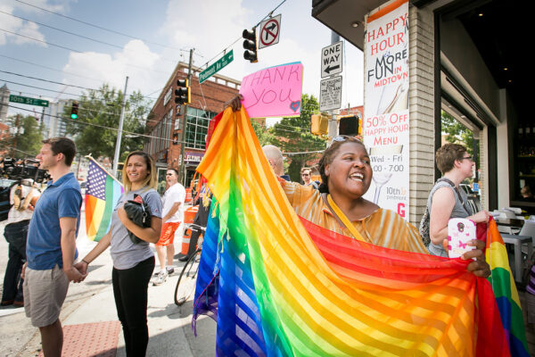 A feminine presenting black person stands near a street corner holding a rainbow flag and a handwritten sign that reads 