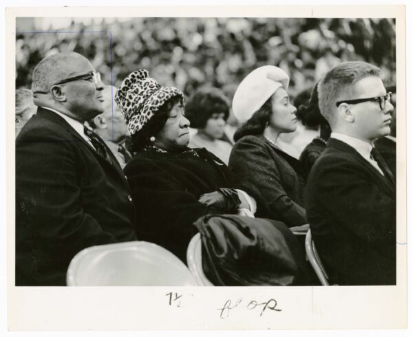 An older Black man in a suit and glasses, an older Black woman wearing a dark jacket and a leopard print hat, and a younger Black woman in a dark jacket and light-colored hat sit outside at an event.