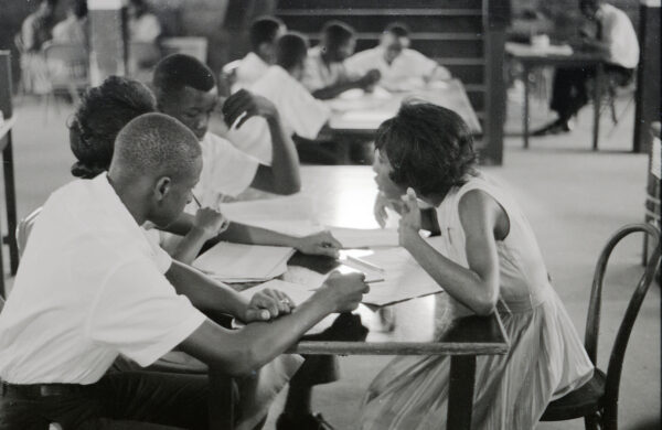 Photograph of four teenaged Freedom School students take part in class discussion sitting at a table in the basement room of probably St. Paul United Methodist Church. Alice Adams is on the right and George Ann Adams on the left between two male students. More students sit at a table in the background, at the foot of a flight of stairs.