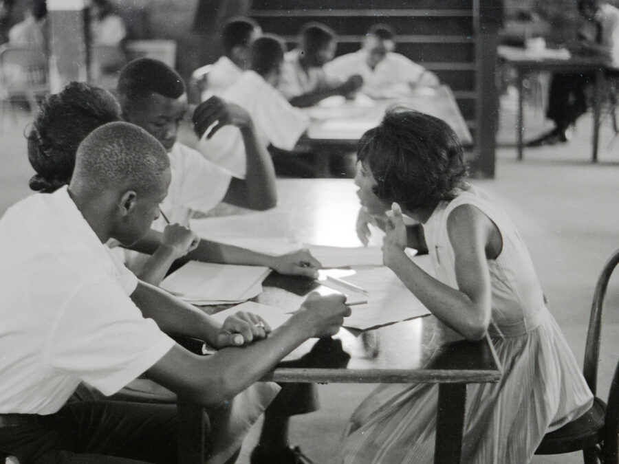 Photograph of four teenaged Freedom School students take part in class discussion sitting at a table in the basement room of probably St. Paul United Methodist Church. Alice Adams is on the right and George Ann Adams on the left between two male students. More students sit at a table in the background, at the foot of a flight of stairs.