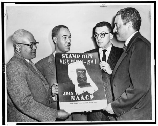 Four Black men wearing suits stand together holding a poster with a dark background and a light colored outline of the state of Mississippi while above the state outline white text reads 