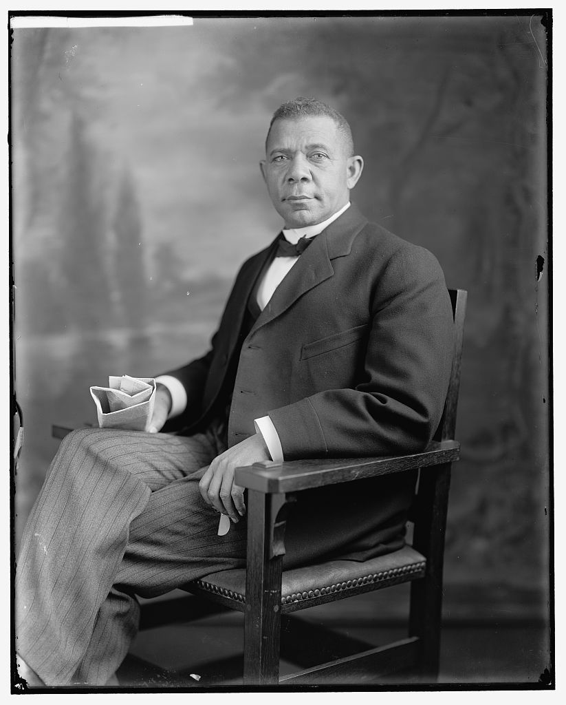 A black and white photograph shows an older Black man wearing a dark suit jacket with a dark bowtie and lighter pinstriped pants sits in a chair in front of a painted background