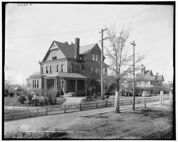 A black and white photo of a large three-story brick home with a neatly manicured lawn and a white picket fence.