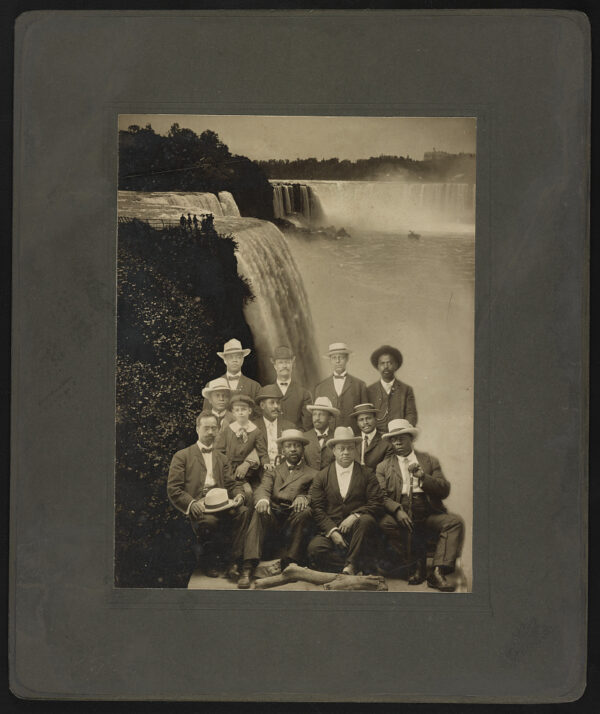 Twelve Black men and one young boy wearing suits and hats pose for a group photo. The photo of the men has been superimposed onto a picture of Niagra Falls.