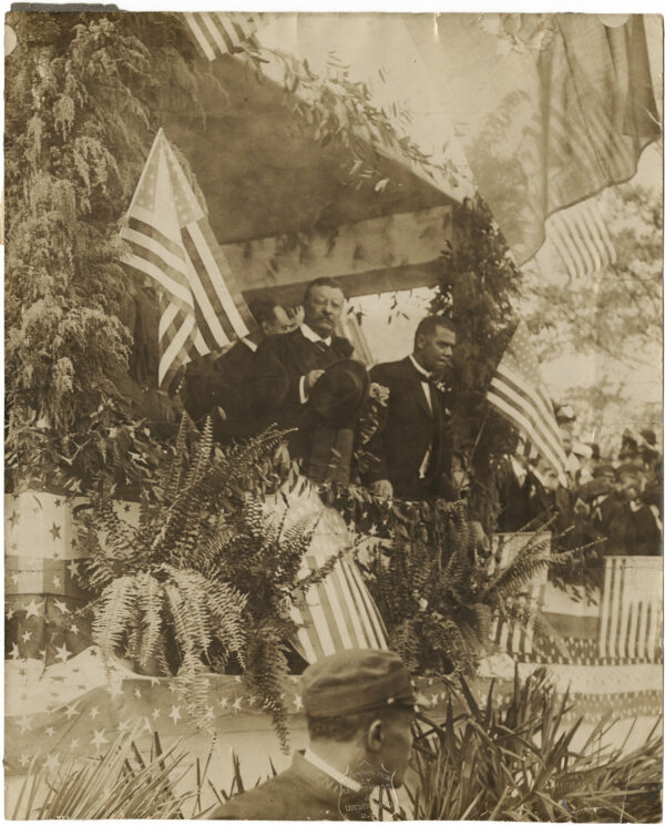 President Theodore Roosevelt stands with hat in hand next to Booker T. Washington on a dais decorated with American flags and greenery.