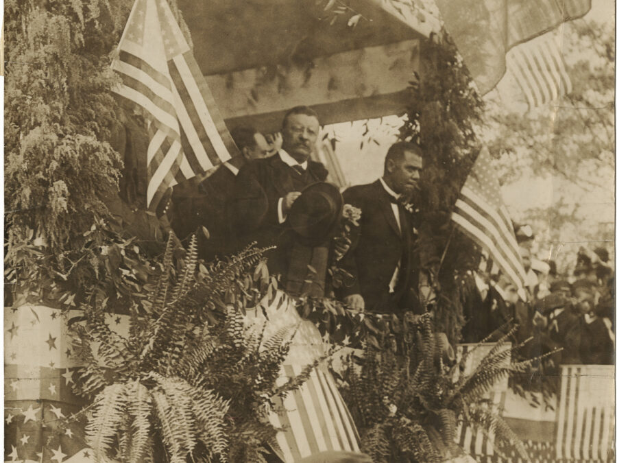 President Theodore Roosevelt stands with hat in hand next to Booker T. Washington on a dais decorated with American flags and greenery.