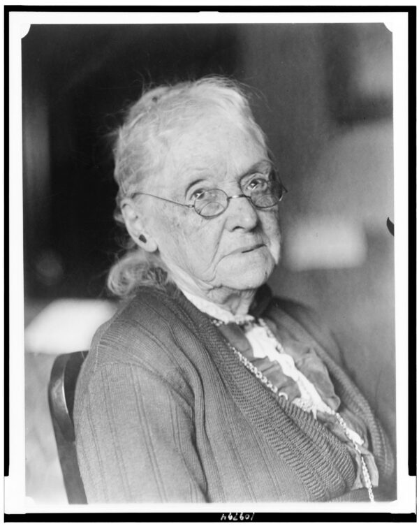 An elderly white woman with small glasses stares into the camera with a serious look.