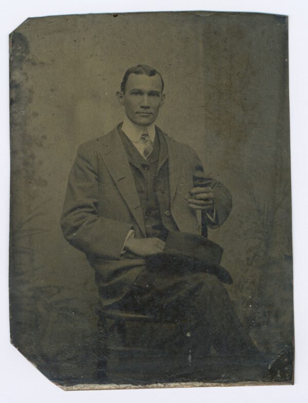 A man wearing a suit with slicked down hair with subtle waves sits on a chair with a hat on his knee.