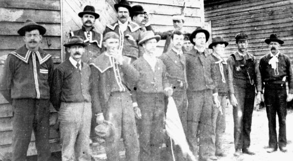 A group of men of varying ages stand in front of a building wearing a variety of mis-matched shirts with light colored trim that seem intended to be a uniform of sorts.