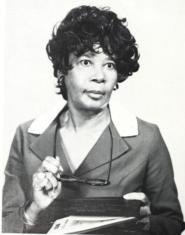 An older Black woman with short layered hair wearing a grey suit jacket with a light colored collar holds a pair of glasses and a book.
