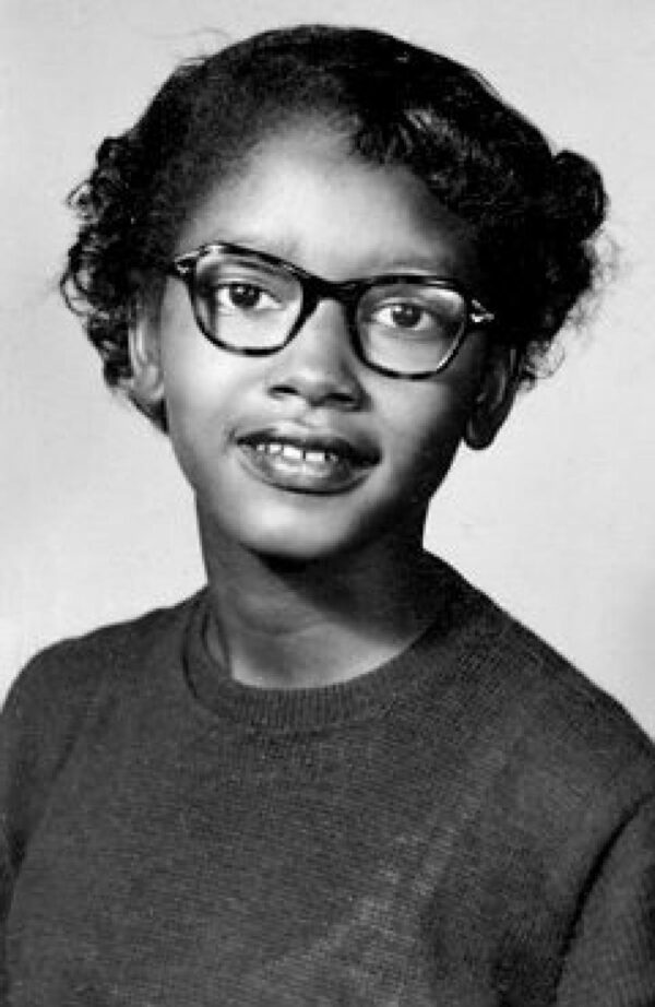 A young Black girl with short hair in a neatly curled hair style is wearing glasses and a plain t-shirt.
