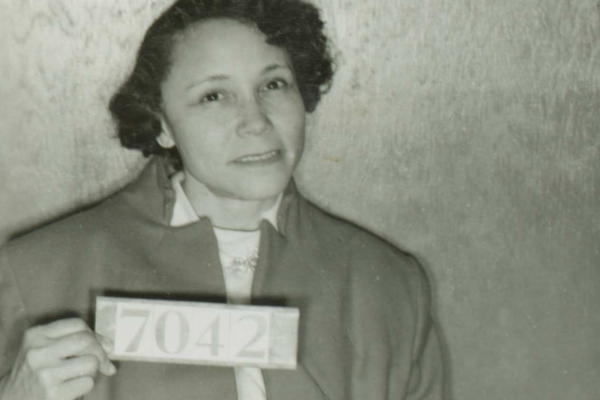 A middle-aged Black woman with short hair in a sleekly curled style wearing a neat cold weather clothes is seated and holding a numerical sign that reads 