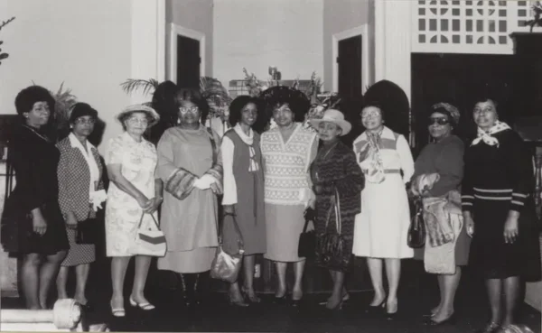 Ten Black women of varying ages wearing their Sunday best pose together at the front of a large room.