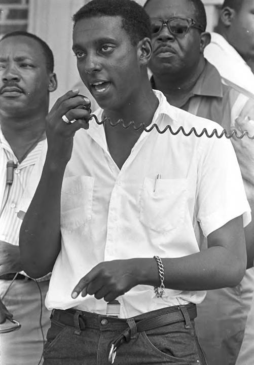 A young Black man wearing a simple white shirt and jeans speaks emphatically into a speaker microphone.