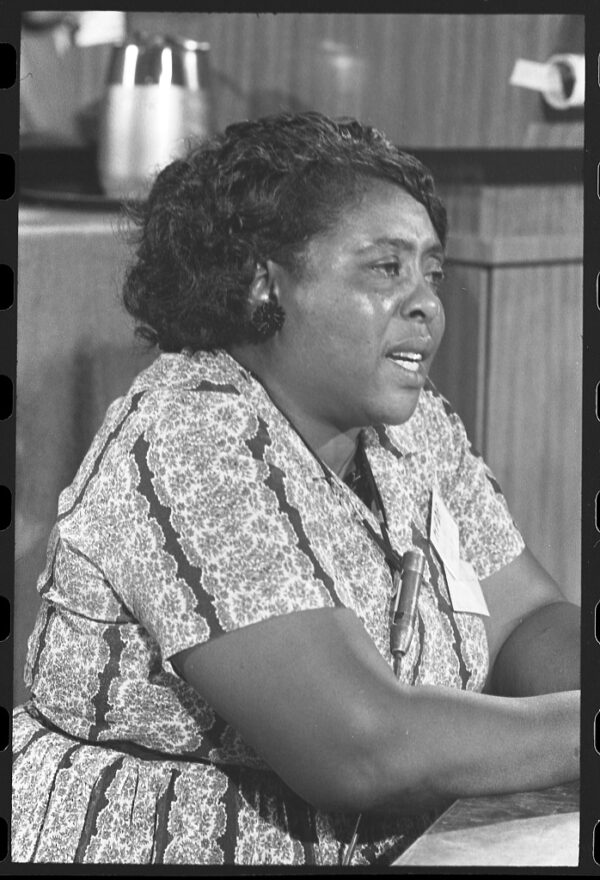 A middle-aged Black woman in a simple patterned dress is sitting at a table and speaking into a microphone.