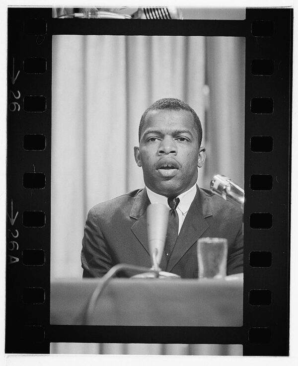 A young Black man wearing a dark suit with a white shirt and dark tie stands at a podium speaking before an audience.