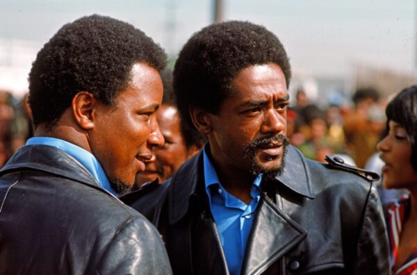 A candid photo of two Black men wearing leather jackets and blue button up shirts standing outside amongst a large crowd of people.