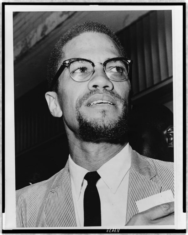 Black and white photo of a Black man with a goatee, moustache, short dark hair with hints of grey wearing glasses with dark rims, a light-colored pin strip suit, a white collared shirt, and a thin black tie.