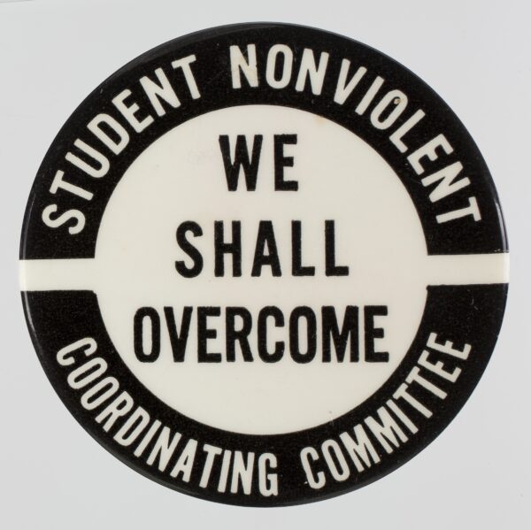 A black and white pin-back button with text. In two arcs around the top and bottom edges of the button face are black blocks with white text. In the middle, centered, is black text. The text reads: [STUDENT NONVIOLENT / WE / SHALL / OVERCOME / COORDINATING / COMMITTEE].