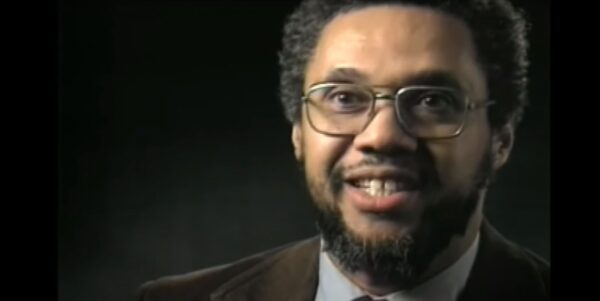 A still frame from an older, lower quality video shows a middle-aged Black man with a beard and short hair wearing a brown suit coat sitting in front of plain dark background.