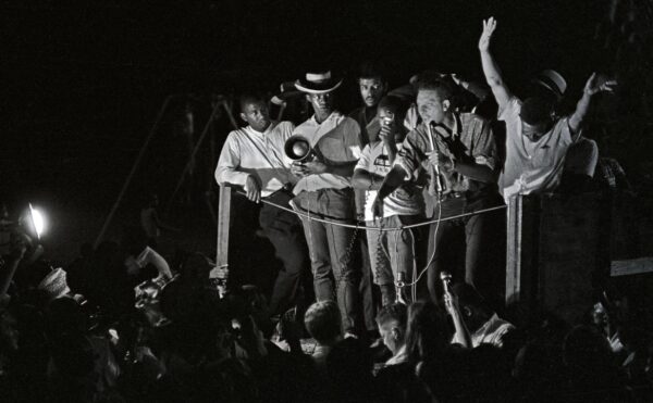 A nightime rally is dramatically lit in this black and white photo that shows a young Black man standing on a stage speaking passionately into a microphone as several other men stand around him holding other microphones and speakers to allow the large crowd to hear the speakers words.