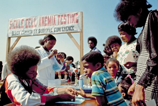 A Black volunteer pricks a Black child's finger as other children and adults stand nearby. A sign behind the volunteers reads 