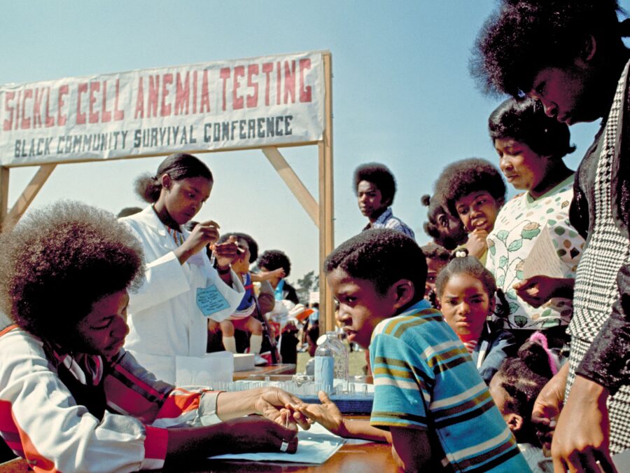 A Black volunteer pricks a Black child's finger as other children and adults stand nearby. A sign behind the volunteers reads 