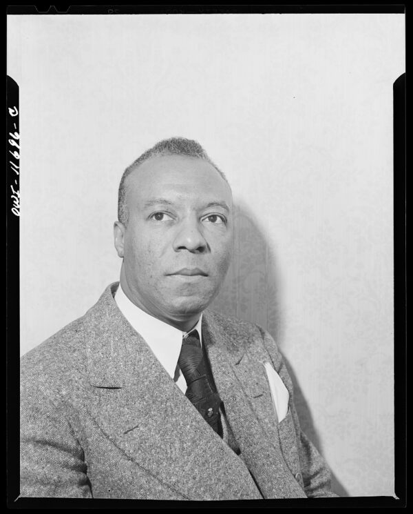 An older Black male with short salt and pepper hair wearing a lighter colored wool suit with a dark colored tie sits for a portrait photo.