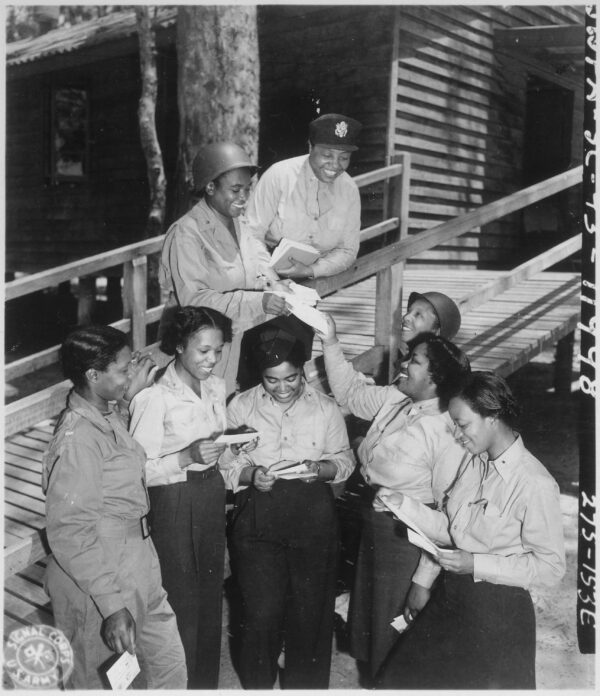 Eight Black women in various different military uniforms smile as they receive and read their mail.