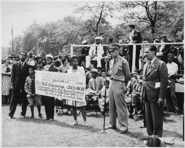 A group of mostly Black men, women, and children are gathered while a soldier speaks at a microphone and a boy and girl hold an over-sized check addressed to the U.S. Treasury for $263,148.83.