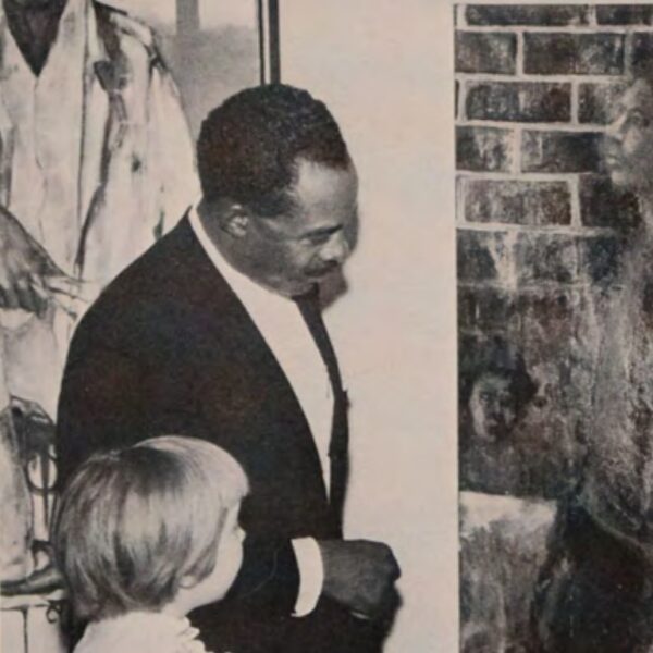 A Black man with short hair wearing a dark suit and white shirt is looking at a piece of art while a small white child stands nearby looking at the same piece.