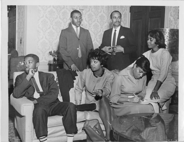 One young Black man and three young Black women sit on a couch while two Black men stand behind. All the men are wearing suits and ties and all the women are wearing professional dresses. One woman takes notes while others seem to be engaged in conversation with someone out of frame.