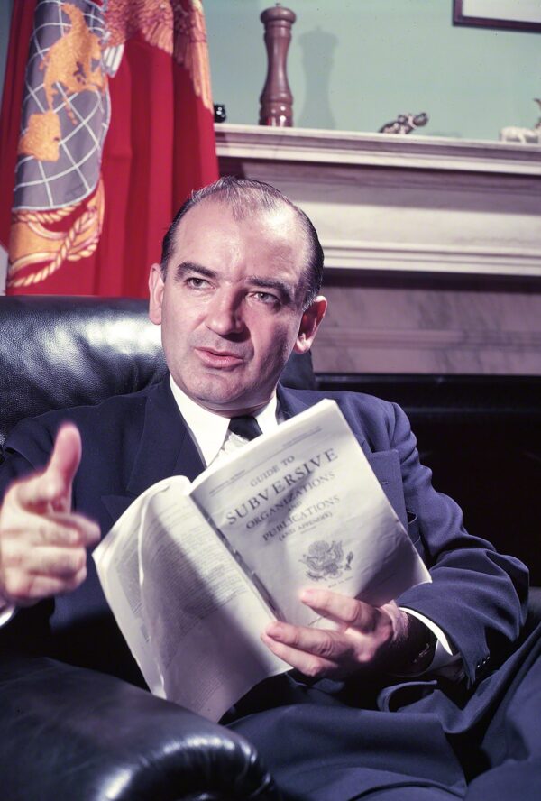 A balding middle-aged white man sits in a leather chair in an office and points towards the photographer while holding a pamphlet titled 