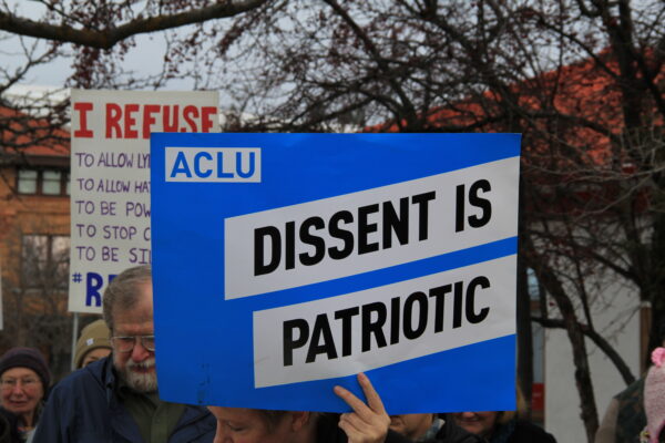 Someone in a group of people outdoors is holding a large blue sign that reads 