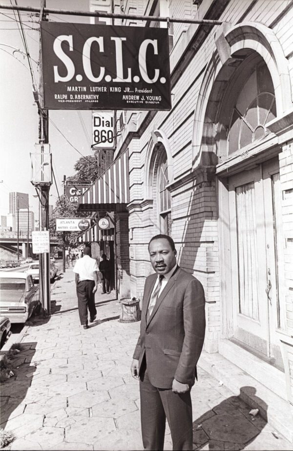 A middle-aged Black man wearing a suit and tie stands on the side walk looking into the camera. Above the man is a sign that reads 