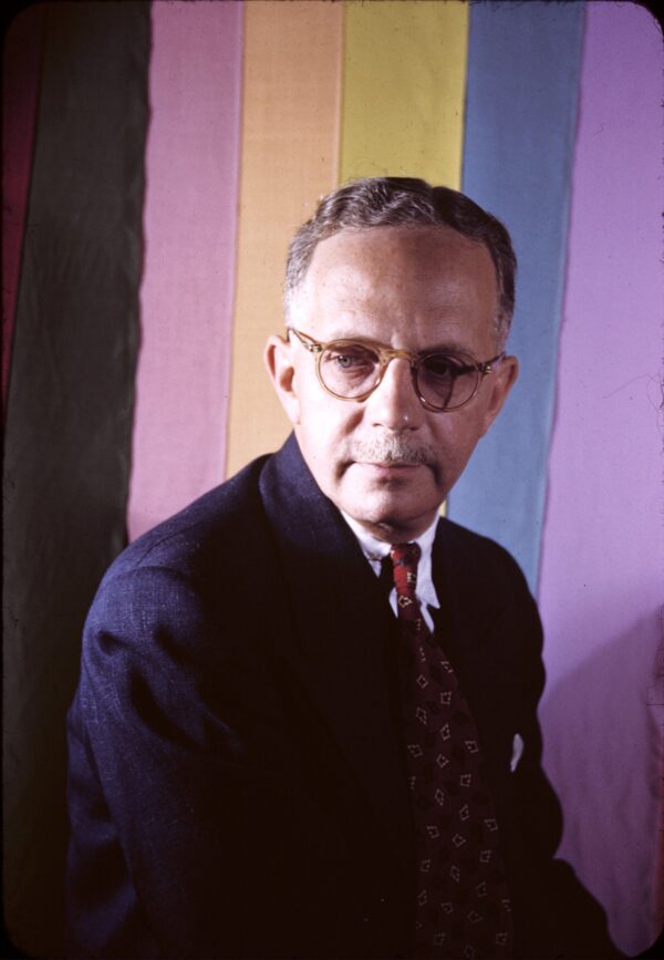 A Black man with very light skin, a grey mustache, and short grey wavy hair sits in front of a colorful background facing the photographer while looking down and to the viewer's left.