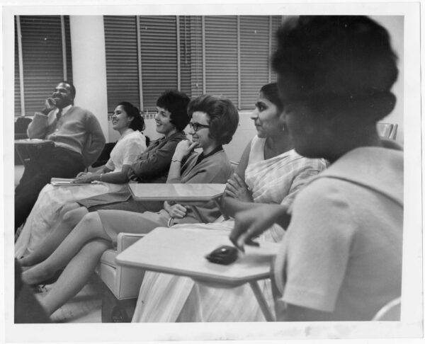 Six students of different races, genders, and ages sit in chairs and at desks as they engage other students off camera in dialogue.