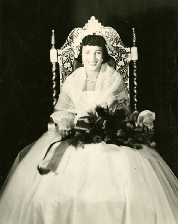 A young Black woman in a ball gown with elegant jewelry and hairdo sits on a throne and holds a large bouquet.