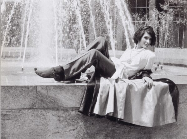 A female-presenting person with short dark hair wearing a tunic and bellbottoms laying in front of a fountain.