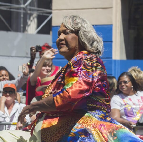 A female-presenting person with grey hair is wearing a multi-colored silk top and riding in the back of a convertible as part of a parade.