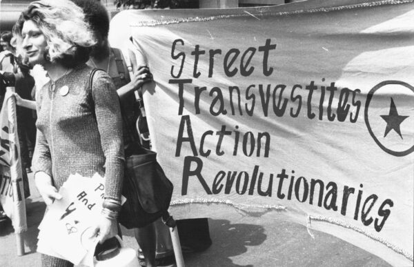 A female-presenting person with light colored hair walking in front of people holding a sign that reads 