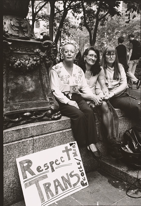 Three female-presenting people sit outside holding hands and looking directly at the photographer.