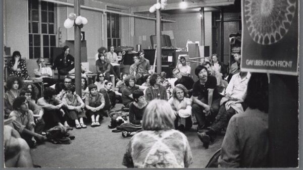 A large group of people sit in a circle on the floor and one chairs and tables in a large open room.