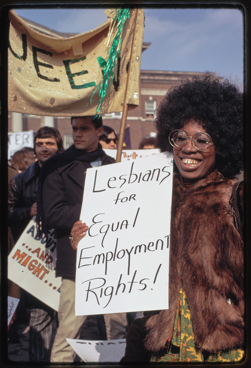 A female-presenting Black person with an afro, large round glasses with a metal rim, and a fur coat holds a sign that reads 