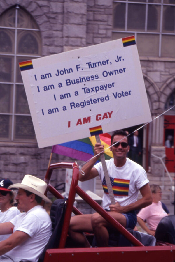 A male-presenting white person sits in the back of a convertible and holds a sign that reads 