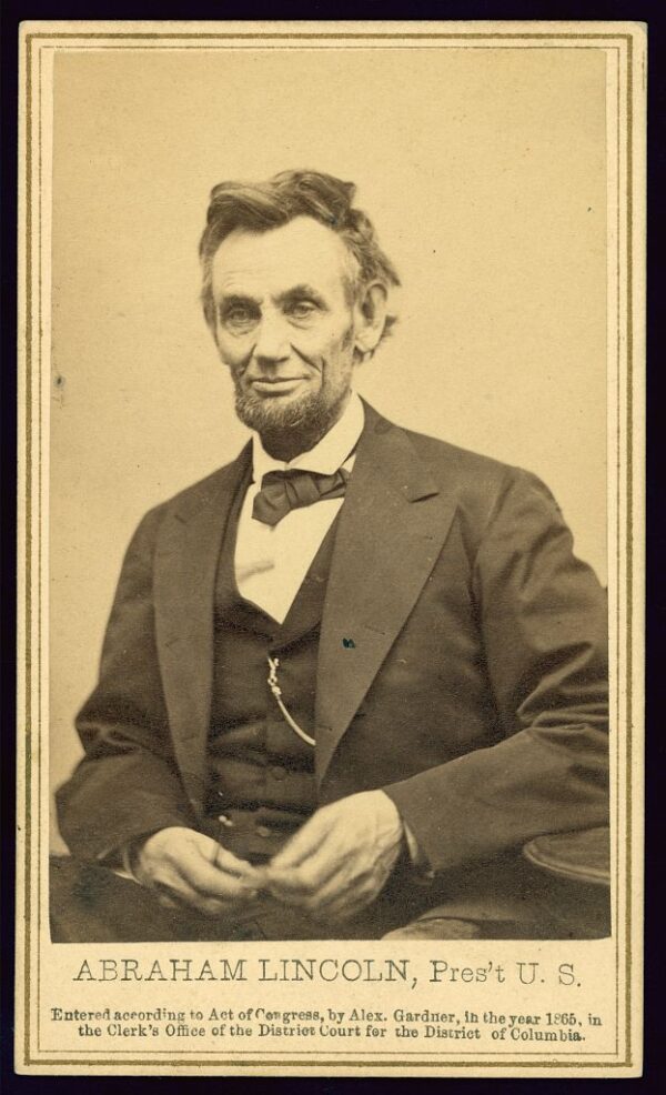 A waist up portrait of an older, thin, white man with deep set eyes and a short salt and pepper beard only on his chin, wearing a dark suit and waist coat.