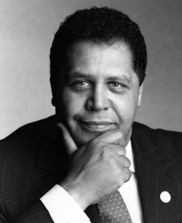 A Black man with dark short hair wearing a dark suit jacket with white shirt and patterned tie looks directly into the camera with his hand on his chin and a slight smile.