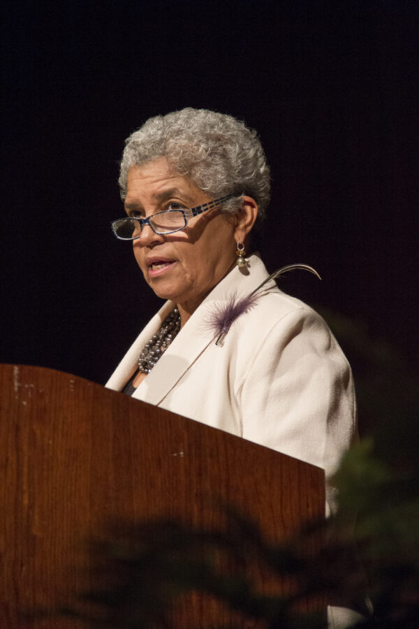 A Black woman with short gray hair wearing glasses and a white suit jacket stands at a podium speaking.