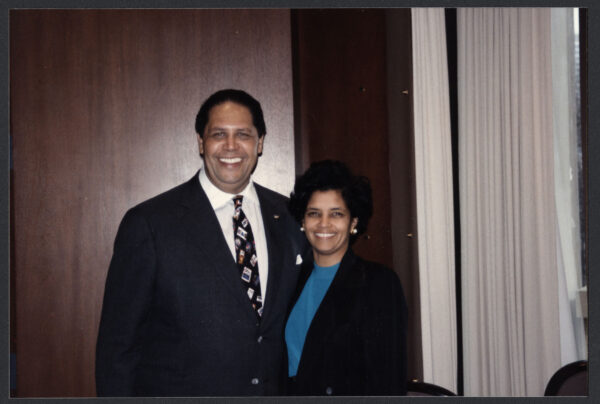 A tall Black man wearing a suit and tie smiles wide while standing next to a shorter Black woman wearing a suit jacket and blue blouse.