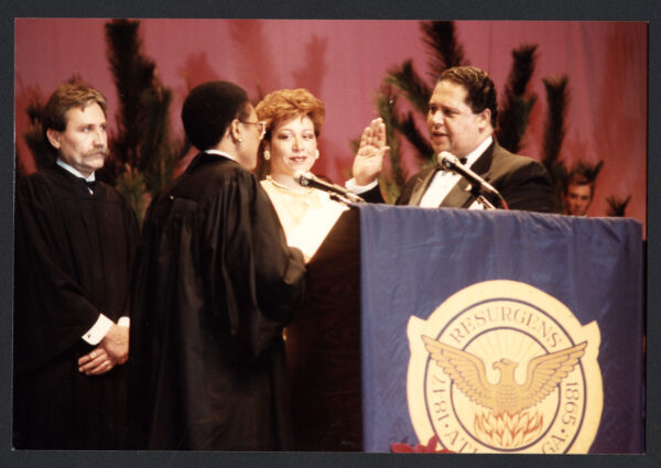 A Black man with short dark hair wearing a black tuxedo stands behind a podium holding his right hand up while facing a female judge in black robes.